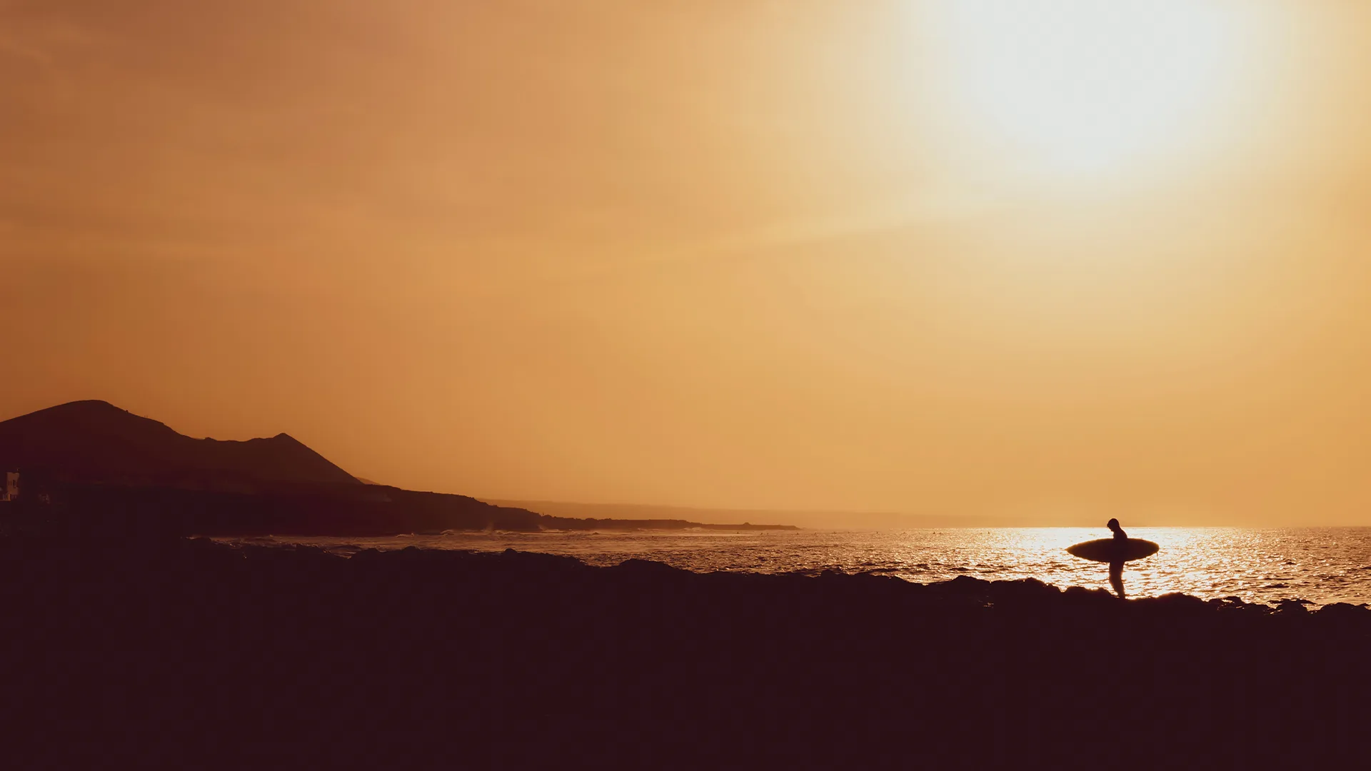 Silhouette of a surfer with a board on the rocks under a burning orange sky