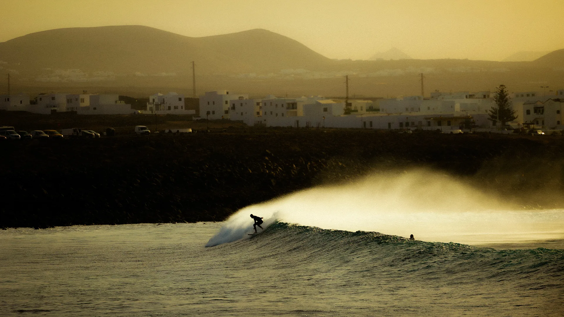 Surfer riding a wind-blown wave at golden hour in front of white houses