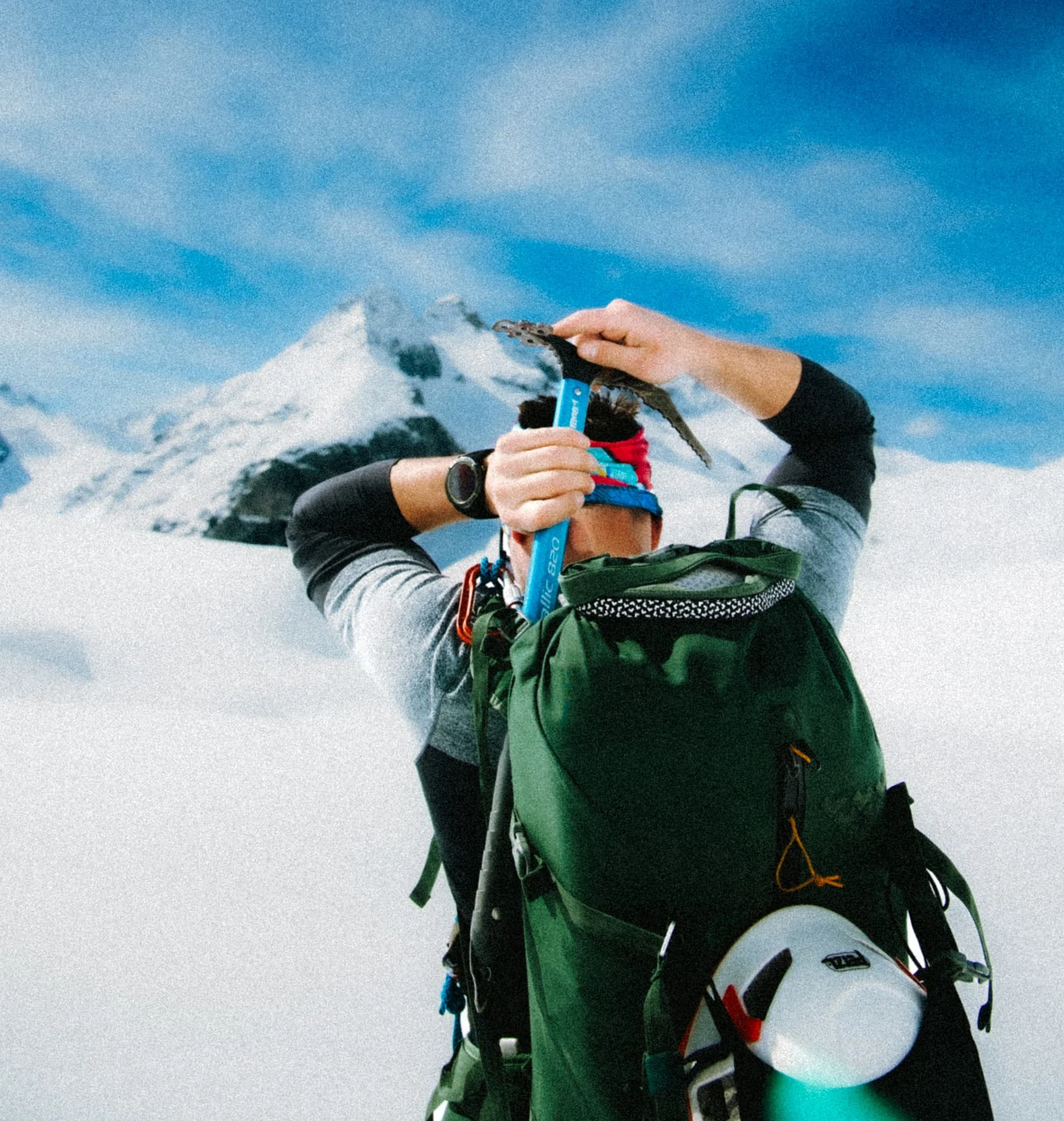 Alpinist with green backpack and ice axe in front of snowy peaks