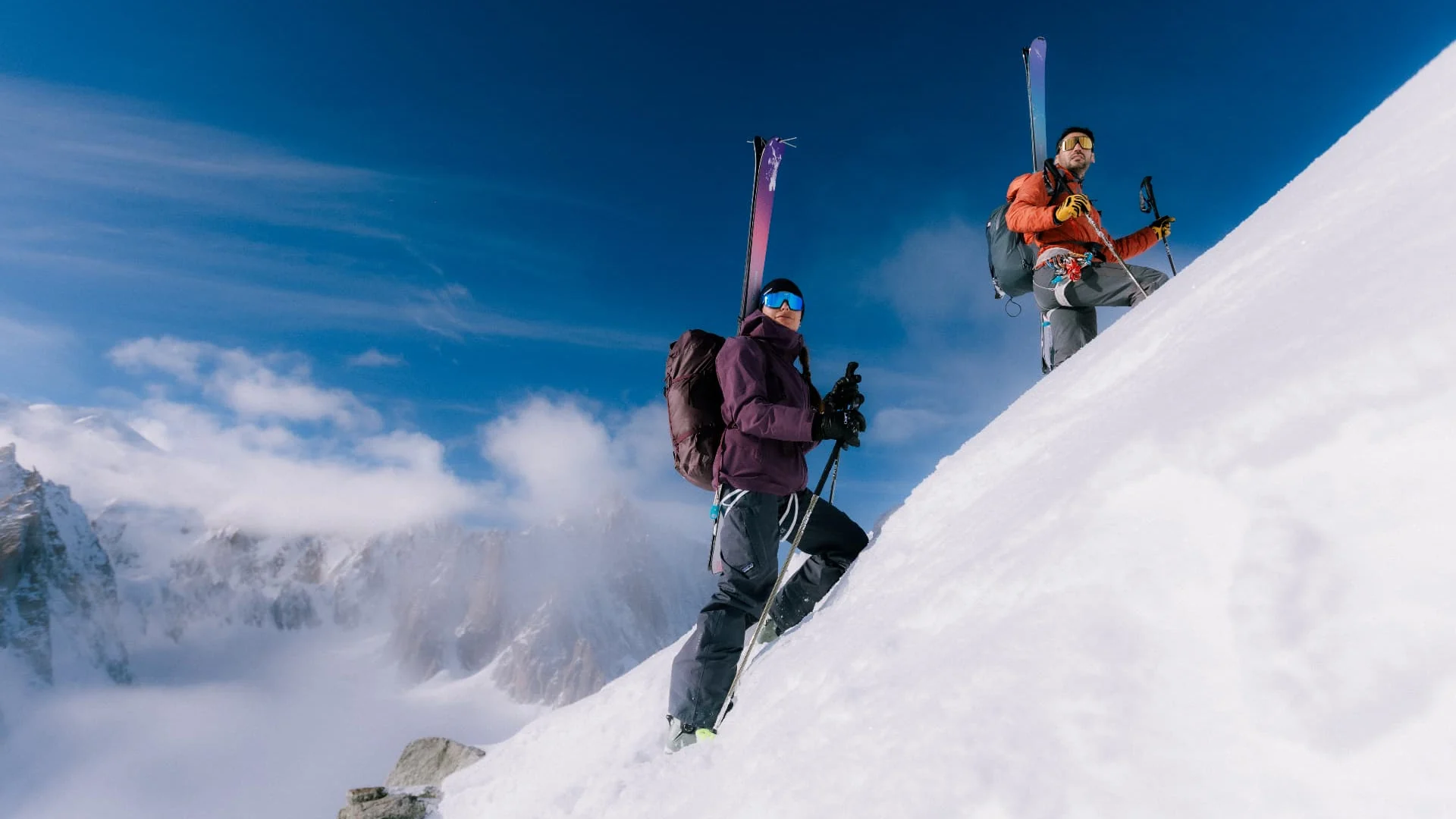 Two ski mountaineers with skis on their backpacks climbing a steep sunlit snowy ridge