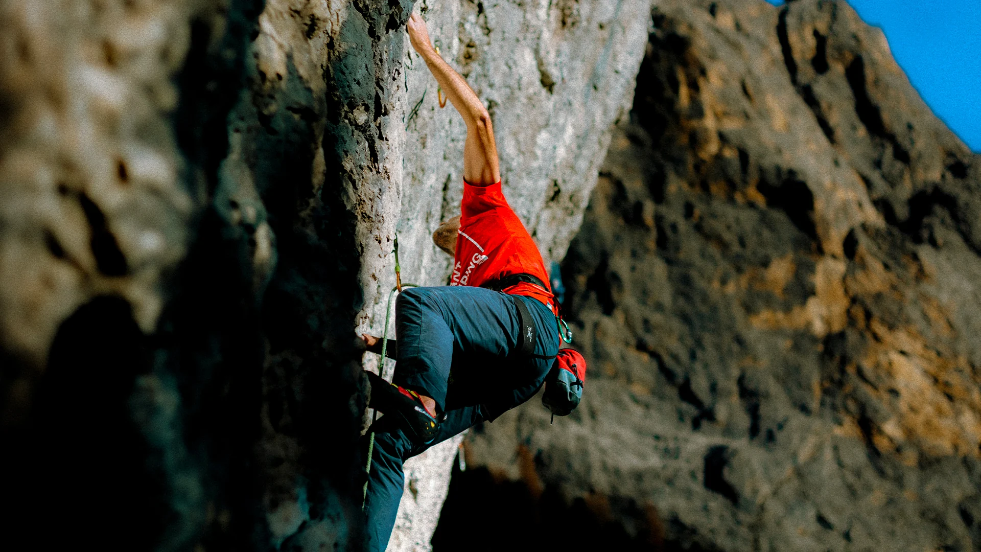 Climber in red ascending a steep rock face beneath a blue sky