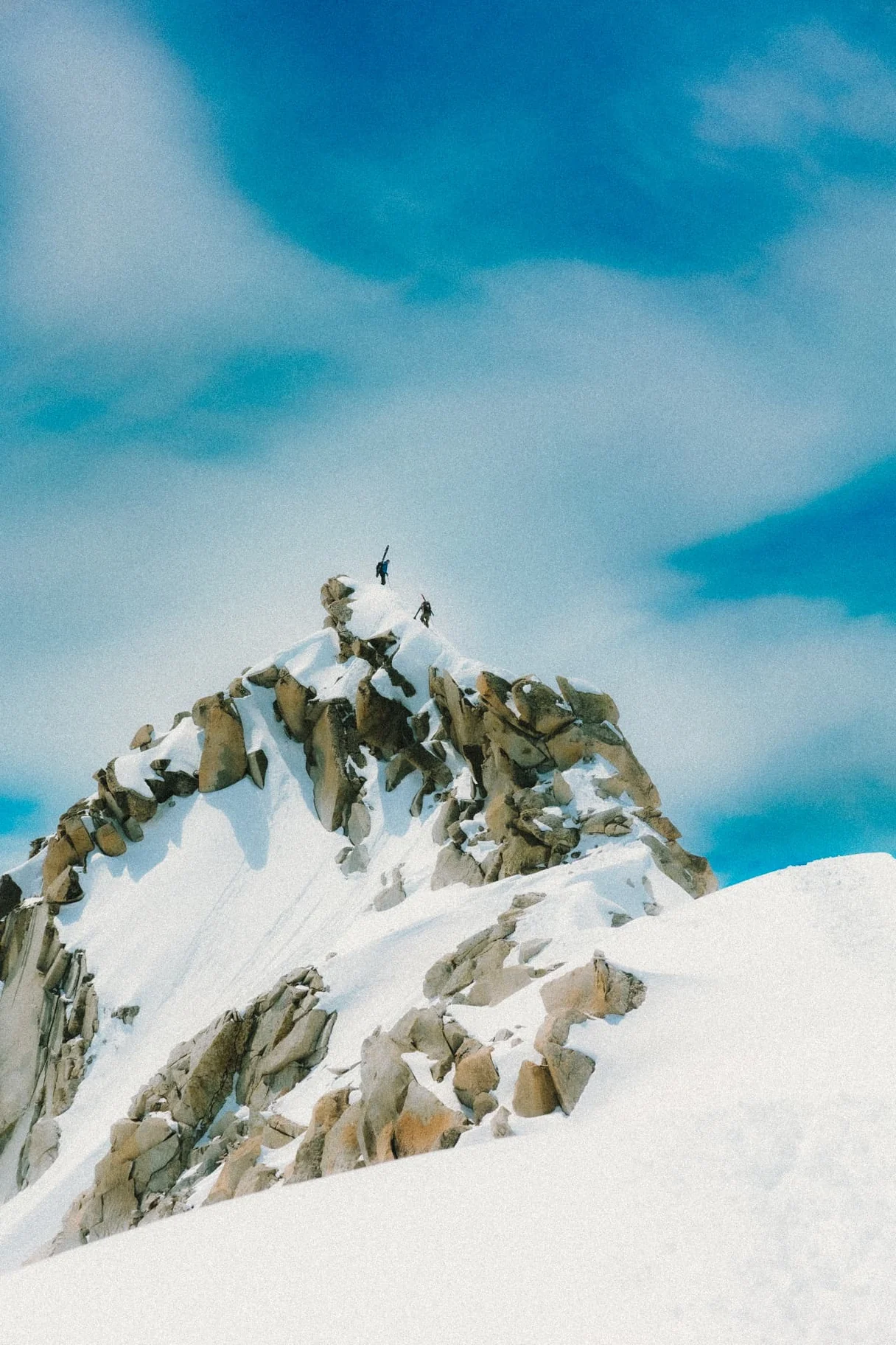 Two ski tourers reaching a rocky summit under a deep blue sky