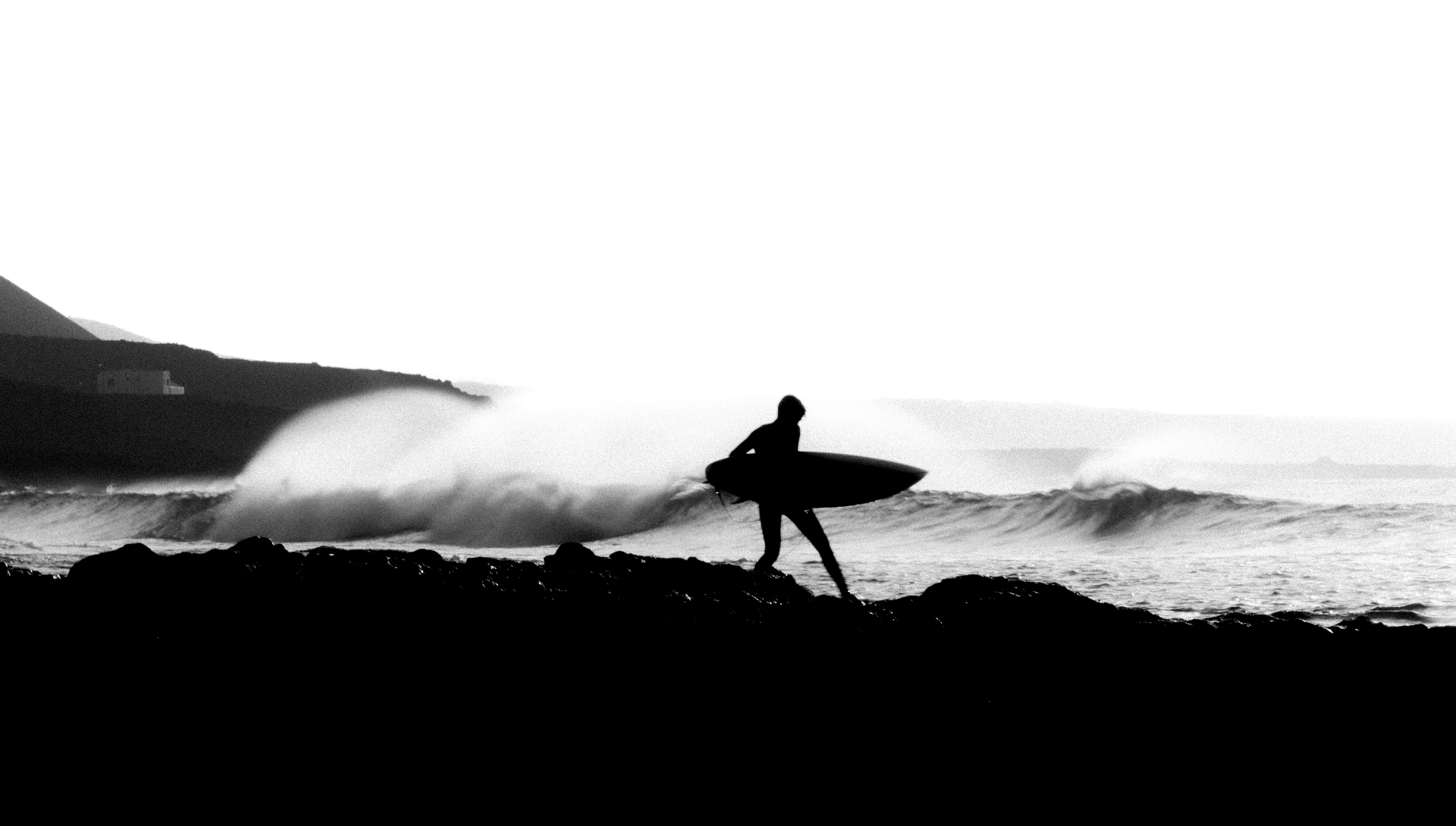 Black-and-white surfer walking over rocks in front of the waves